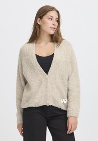 Young woman wearing a beige textured button-up cardigan over a black top and black pants, looking to her left against a white background.