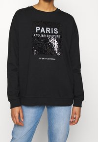 Sudadera negra con un gráfico de lentejuelas que dice "PARIS ATELIER COUTURE" en plata y blanco, con puños y dobladillo acanalados.