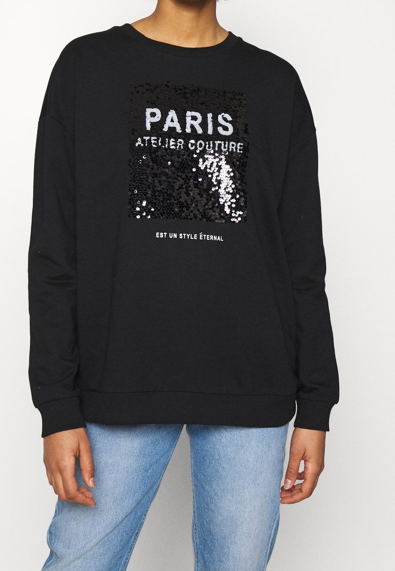 Sudadera negra con un gráfico de lentejuelas que dice "PARIS ATELIER COUTURE" en plata y blanco, con puños y dobladillo acanalados.