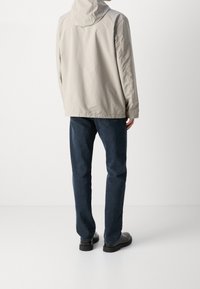 Mężczyzna w beżowej kurtce z kapturem, ciemnoniebieskich dżinsach i czarnych butach stojący tyłem na białym tle.