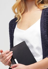 Cárdigan texturizado en azul marino sobre una camiseta de cuello en V blanca, sosteniendo un cuaderno negro con un acento plateado. Diseño simple y moderno con líneas limpias.