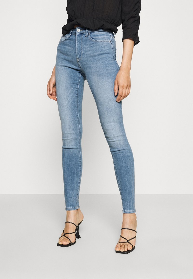 Světle modré skinny džíny vyrobené z denimu, s mid-rise pasem a jemným vyblednutím. V páru s černými sandály na podpatku. Neutrální pozadí.