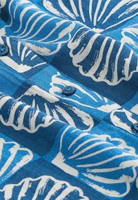 Blue buttoned shirt featuring a shell pattern in white. The fabric has a smooth texture with visible stitching and contrasting blue buttons.