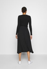 Robe noire à manches longues avec une taille cintrée, une silhouette fluide et des bords de manches effilochés, associée à des baskets blanches.