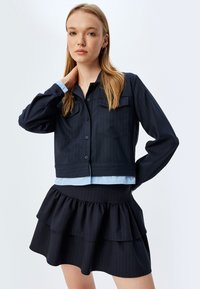 Giacca accorciata a righe navy con due tasche sul petto, indossata sopra una camicia azzurra, abbinata a una gonna nera a strati con volant.