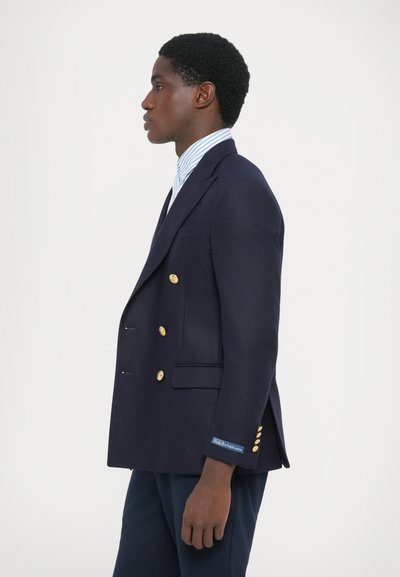 Blazer bleu marine à double boutonnage avec des boutons dorés, poches latérales et coupe ajustée, associé à une chemise rayée bleu clair en dessous.