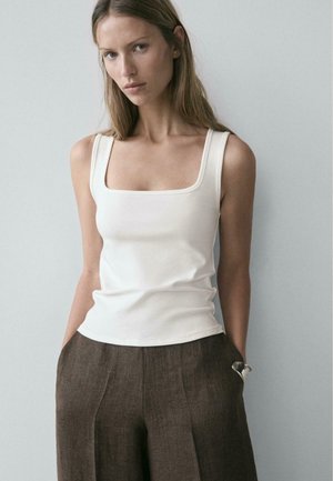 Donna con lunghi capelli chiari che indossa una canotta bianca con scollo quadrato e pantaloni marroni a vita alta, su uno sfondo neutro.