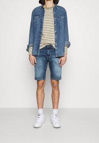 Mies, joka on pukeutunut siniseen farkkupaitaan beigen ja valkoisen raidallisen pitkähihasen paidan päällä, farkkushortseihin, valkoisiin sukkiin ja valkoisiin lenkkitossuihin, seisoo yksinkertaista taustaa vasten.