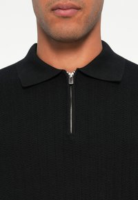 Jersey negro de cuello con cremallera. Presenta un patrón texturizado y un detalle de cremallera metálica en el escote.
