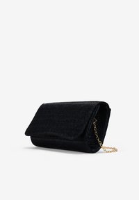 Bolso de mano negro hecho de tela texturizada, con un cierre de solapa y una correa de cadena dorada desmontable. Forma compacta y rectangular.