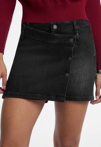 Crna traper suknja s dizajnom omota, s pet dugmadi sprijeda, izgužvanim rubom i malo teksturiranim materijalom.