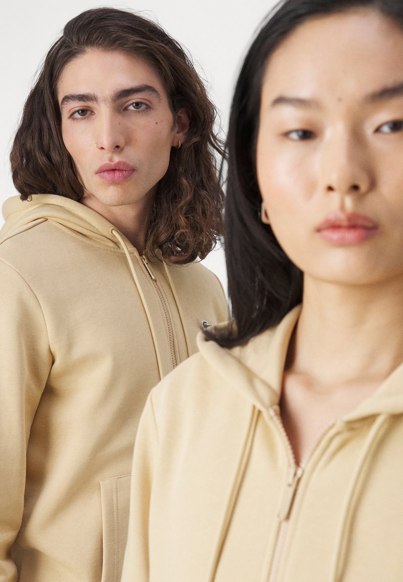 Beige zip-up hoodies met kangoeroezakken, gemaakt van zacht stof en met een subtiele branding op de linkerborst.