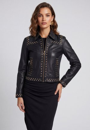 Frau trägt eine schwarze Lederjacke mit goldenen Nieten und Reißverschlüssen, kombiniert mit einem schwarzen, eng anliegenden Rock, und posiert vor einem einfarbigen Hintergrund.