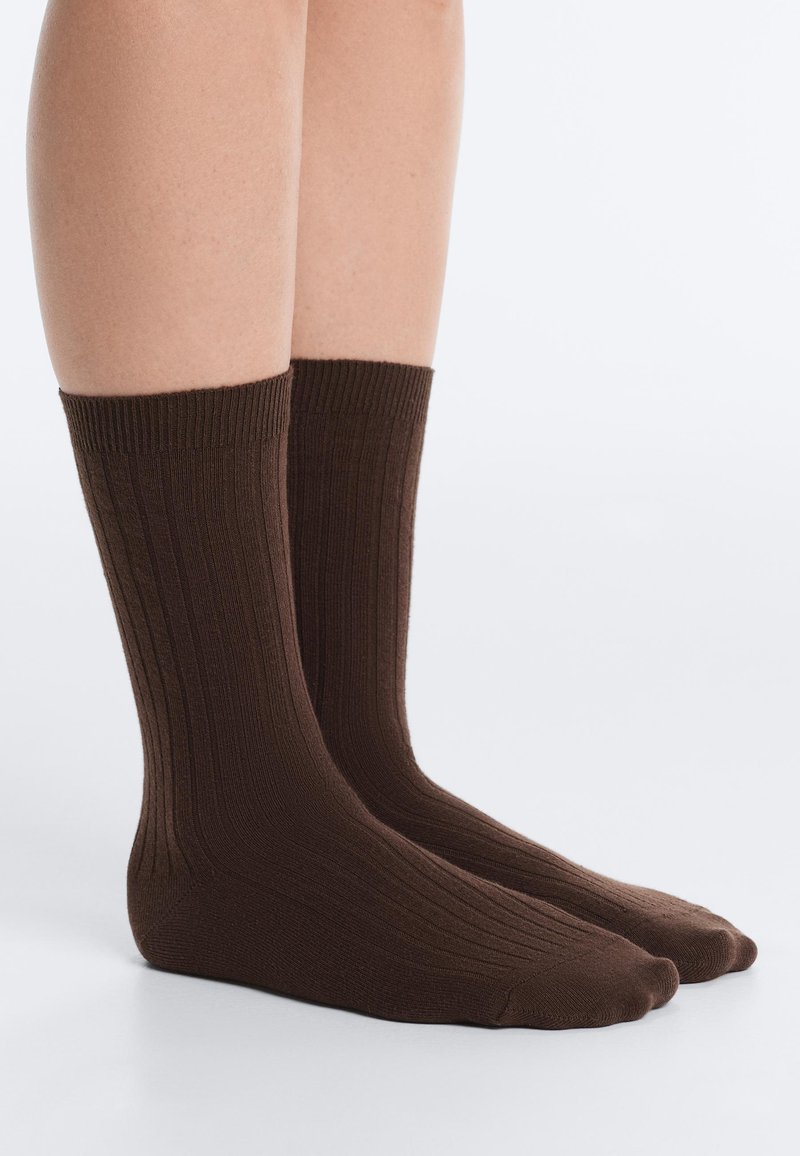 Jambes portant des chaussettes côtelées brun foncé mi-mollet, debout sur une surface lisse et claire.