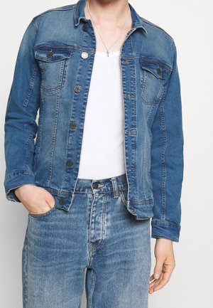 Persona vestida con una chaqueta de mezclilla azul y jeans sobre una camiseta sin mangas blanca, con un collar de cruz de plata y una mano en el bolsillo.