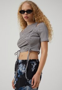 Sivi rebrasti crop top s naboranim dizajnom, kratkim rukavima i podesivim vezicama. Uparen s crnom suknjom s cvjetnim uzorkom.