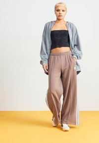 Siva zip-up dukserica preko črne kratke majice, v kombinaciji z ohlapnimi rjavimi trenirkami z belimi črtami in belimisupergami. Gladka tekstura tkanine.