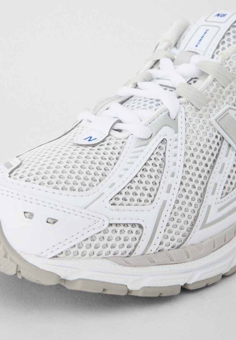 Scarpa sportiva bianca e grigia con tomaia in mesh, chiusura con lacci e suola testurizzata con un evidente logo "N" e sottili dettagli di design.