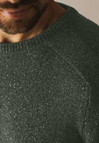 Donkergroen gebreide trui met een textuur en lichte spikkels, ronde halslijn en raglanmouwen.