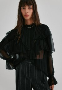 Černá průsvitná blůza s vrstvenými volány, vysokým límcem a dlouhými rukávy se volánovými manžetami, spárovaná s pruhovanými tmavými kalhotami.