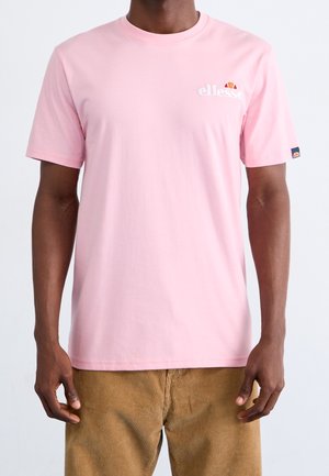 Mann trägt hellrosa Ellesse-T-Shirt und braune Cordhosen, steht mit entspannten Armen an den Seiten vor einfachem Hintergrund.