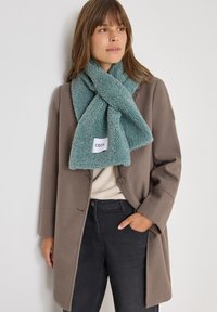 Faux fur halstørklæde i blød teal med en tekstureret, fluffy overflade. Label synlig med mærkenavnet "CECIL." Sammen med en brun frakke.