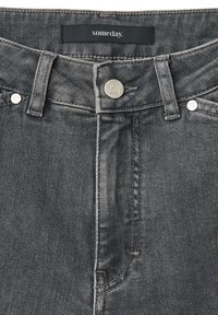 Close-up van de tailleband van een grijze spijkerbroek met een metalen knoop en klinknagels, met een zwart label waarop het woord "someday" staat.