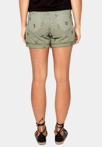 Shorts brodés vert olive avec ourlets retroussés, présentant des motifs floraux et deux poches arrière, associés à des espadrilles noires.