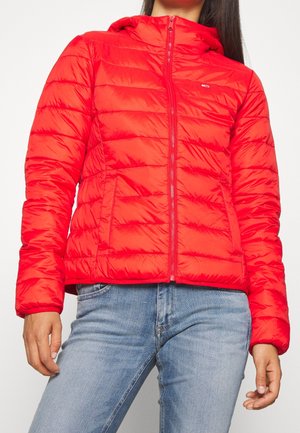 Kobieta w jasnoczerwonej pikowanej kurtce z przednim zamkiem błyskawicznym, w połączeniu z jasnoniebieskimi dżinsami, stoi na jednolitym tle.