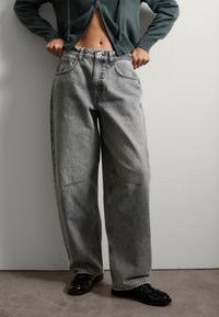 Vaqueros de pierna ancha gris claro con cintura alta, que presentan grandes bolsillos frontales y sutiles detalles de costura. Combinados con zapatos negros brillantes.