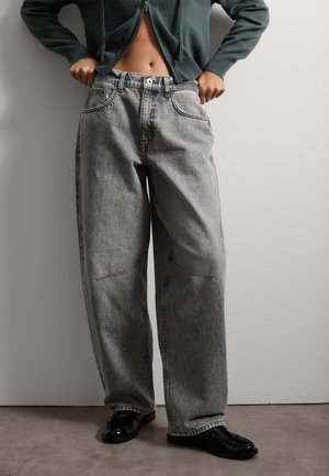 Vaqueros de pierna ancha gris claro con cintura alta, que presentan grandes bolsillos frontales y sutiles detalles de costura. Combinados con zapatos negros brillantes.