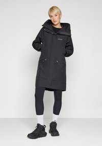 Svart vanntett parkas med hette, med glidelåslommer, rett snitt og glatt tekstur. Kombinert med mørke leggings og svarte sko.