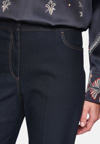 Dark blue denim trousers with visible stitching, featuring a side pocket and a smooth texture. A patterned blouse is partially visible above.