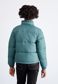 Zeleno-modra puhovka z visokim ovratnikom, elastičnimi manšetami in vodoravnimi šivi. Tkanina ima gladko teksturo in lahko zasnovo.