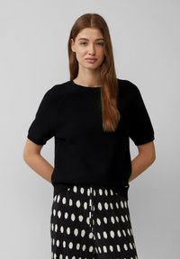 Czarny, dziany sweter z krótkimi rękawami i okrągłym dekoltem. Noszony z plisowaną spódnicą w czarno-białe kropki. Gładka tekstura, luźny krój.