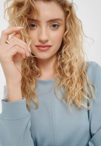 Top azzurro chiaro a maniche lunghe con scollatura rotonda e una texture liscia. La modella ha capelli biondi e mossi e tiene una ciocca in mano, mostrando un anello d'oro.