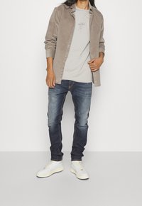 Beige kordflor tröja över en grå T-shirt, i kombination med mörkblå jeans och vita sneakers. Avslappnad, lager-på-lager outfit med enkla designelement.