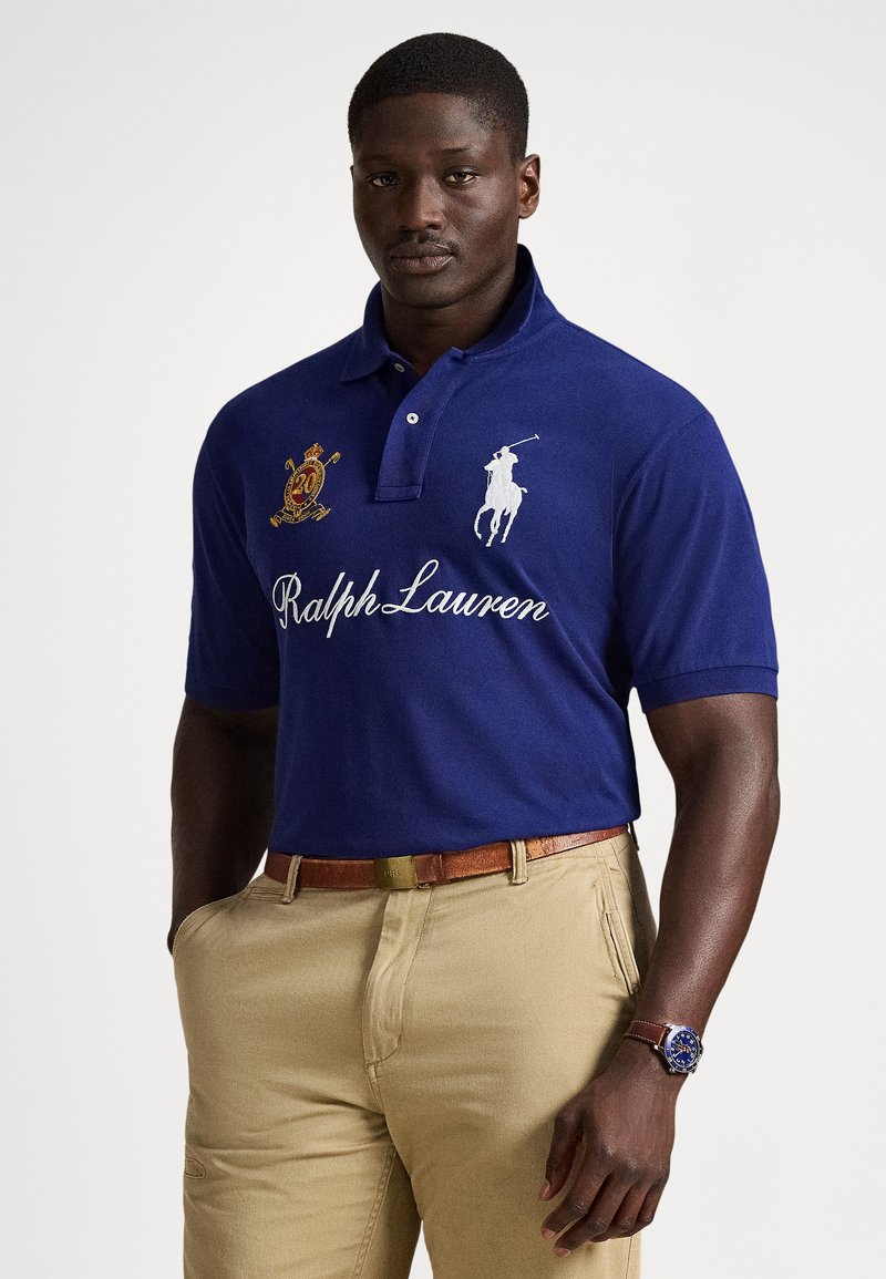 Mann iført blå Ralph Lauren polo skjorte med emblem, beige bukser, brunt skinnbelte og klokke med blå urskive, stående med hendene i lommene.