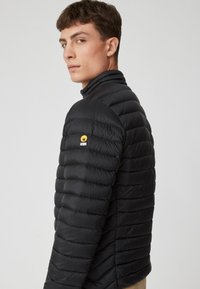 Giacca a vento nera con design trapuntato orizzontale, colletto alto e un patch con logo giallo sulla spalla sinistra. Materiale liscio e leggero.