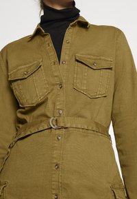 Abito camicia verde oliva con vita con cintura, due tasche sul petto, chiusura a bottoni e colletto. Realizzato in tessuto morbido e resistente.