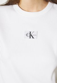 Bílá žebrovaná mikina s kulatým výstřihem, na přední straně s obdélníkovou nálepkou Calvin Klein Jeans. Látka má hladký povrch.