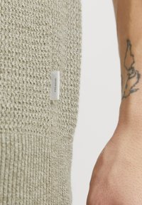 Suéter tejido de color verde claro que muestra un patrón de punto texturizado. Presenta una pequeña etiqueta blanca con la palabra "Premium" en el lateral. Brazo con tatuaje visible.