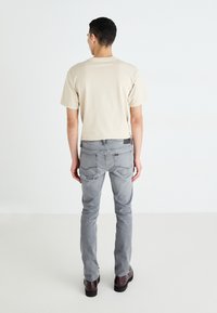 T-shirt en coton beige et jeans en denim gris. Les jeans ont une coupe ajustée et des poches arrière. Le modèle porte des bottes noires et se tient devant un fond uni.
