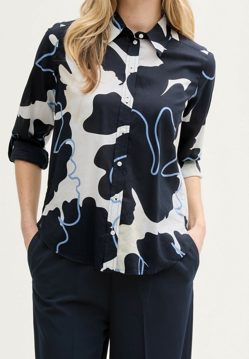 Femme portant une chemise boutonnée à manches longues avec un motif abstrait noir, blanc et bleu, les mains dans les poches et un pantalon foncé.