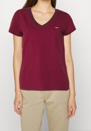 Kvinde iført en kortærmet, mørkerød striped V-hals t-shirt med et lille Levi's logo på brystet og beige bukser.
