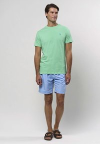 Zelené bavlnené tričko, modré šortky a hnedé sandále. Tričko má malú logo; šortky sú ľahké a dĺžkou po kolená.