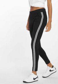 Leggings noirs en matériau extensible, avec deux bandes latérales blanches et une coupe ajustée. Portés avec des baskets blanches.