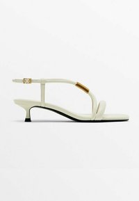 Sandale blanche à talon bas avec des fines lanières, une petite boucle dorée sur la bride de cheville et un accent doré sur la lanière supérieure.
