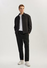 Zwarte jas met twee borstzakken, gedragen over een wit t-shirt, gecombineerd met zwarte broek en witte sneakers. Model staat tegen een neutrale achtergrond.