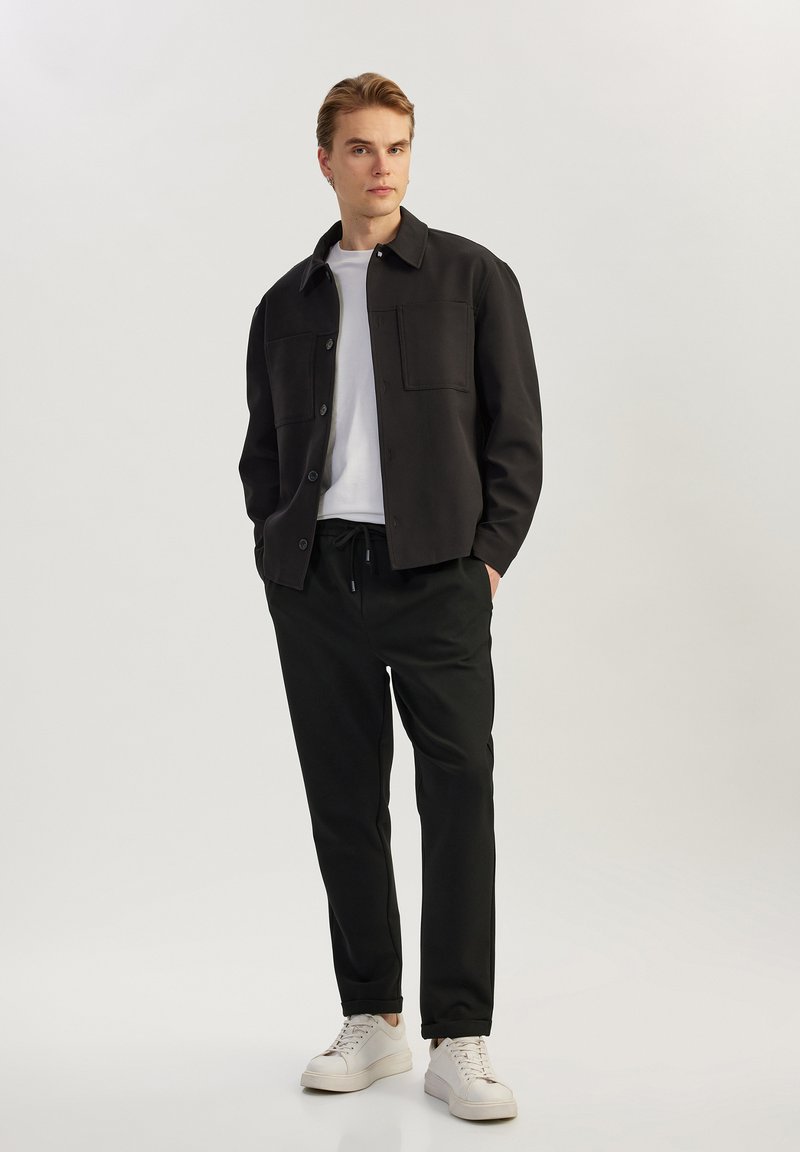 Zwarte jas met twee borstzakken, gedragen over een wit t-shirt, gecombineerd met zwarte broek en witte sneakers. Model staat tegen een neutrale achtergrond.